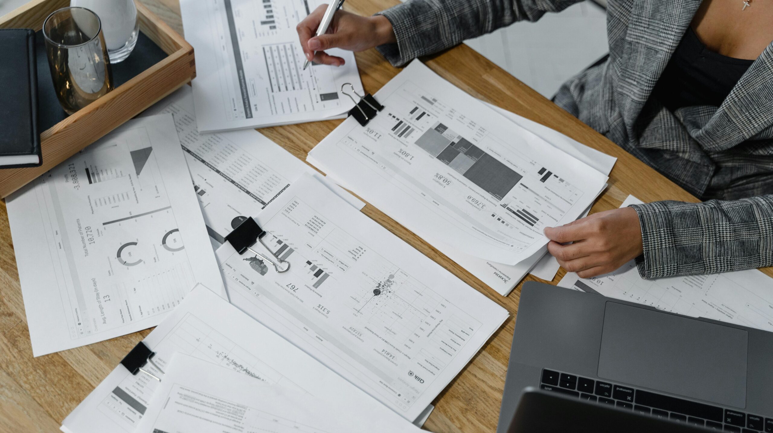 Woman analyzing financial data and reports on a wooden desk with a laptop, showcasing analytics in a workspace.